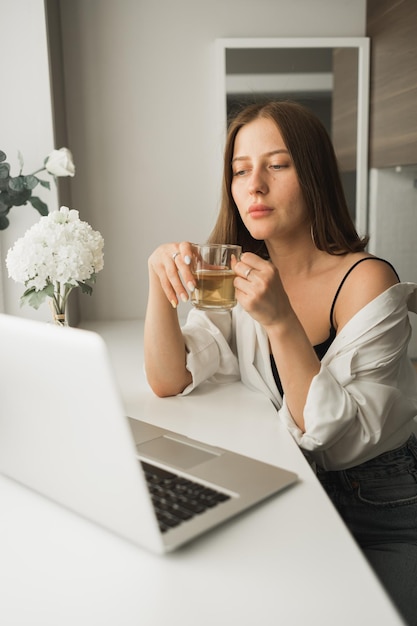 Gros plan sur une jeune jolie femme buvant du thé pendant la pause après avoir tapé et navigué en ligne sur un ordinateur portable w