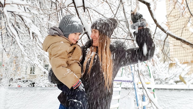 Gros Plan D'une Jeune Mère Joyeuse Avec Son Joli Fils En Veste Et Chapeau Jouant Avec Un Arbre Couvert De Neige Sur L'aire De Jeux Dans Le Parc