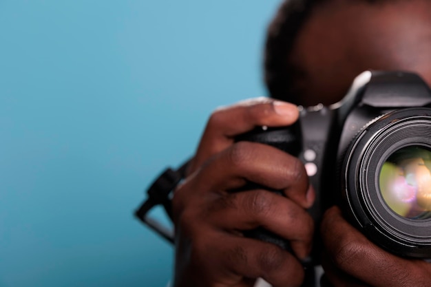 Gros plan d'un jeune photographe afro-américain prenant une photo avec un appareil photo reflex numérique professionnel en se tenant debout sur fond bleu. Passionné de photographie avec appareil photo moderne prenant des photos.