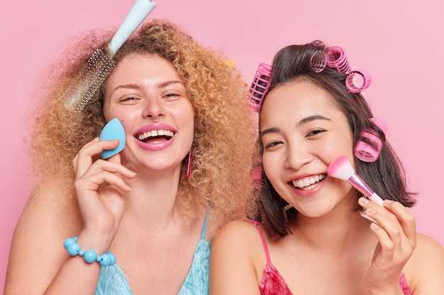 Gros plan de jeunes femmes métisses gaies appliquent un fond de teint ou de la poudre sur le visage avec une éponge et un pinceau cosmétique sourire font largement la coiffure soin de la beauté isolée sur fond rose