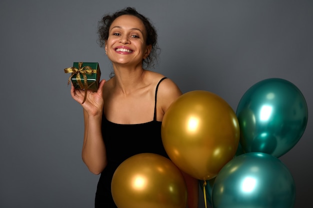 Gros plan d'une jolie femme avec un beau sourire posant avec un cadeau de Noël dans du papier d'emballage brillant et un arc doré, tenant de beaux ballons à air gonflés festifs sur fond gris avec espace publicitaire pour copie