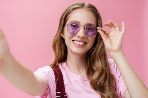 Gros plan d'une jolie femme féminine avec un tatouage de cheveux ondulés et une petite cicatrice sur le bras touchant des lunettes de soleil vérifiant les cadres sur le visage tout en prenant un selfie tirant le bras vers la pose sur fond rose.