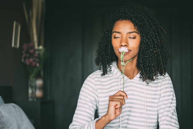 Gros plan d'une jolie femme noire aux cheveux bouclés sentant une fleur rose s'asseoir sur le lit