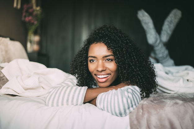 Gros plan d'une jolie femme noire aux cheveux bouclés souriant et allongé sur le lit en regardant loin