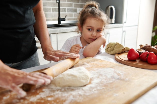 Gros plan sur une jolie fille regardant sa mère dérouler la pâte avec un rouleau à pâtisserie en bois. Maman et sa fille cuisinent des pizzas ensemble.