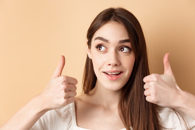 Gros plan d'une jolie fille regarde le logo du coin supérieur gauche, montrant les pouces vers le haut en approbation, comme un produit, debout sur fond beige.