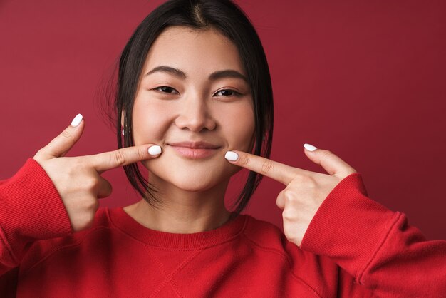 Gros plan d'une jolie jeune femme asiatique souriante portant des vêtements décontractés, isolée sur un mur rouge, tenant les bras au visage