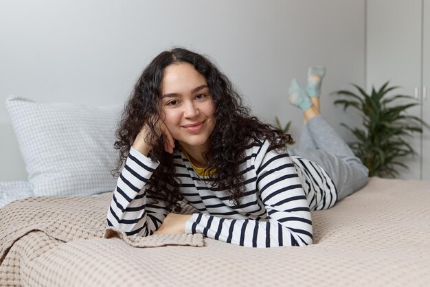 Photo gros plan d'une jolie jeune femme aux cheveux bouclés regardant la caméra se reposer et allongée sur le lit à la maison.