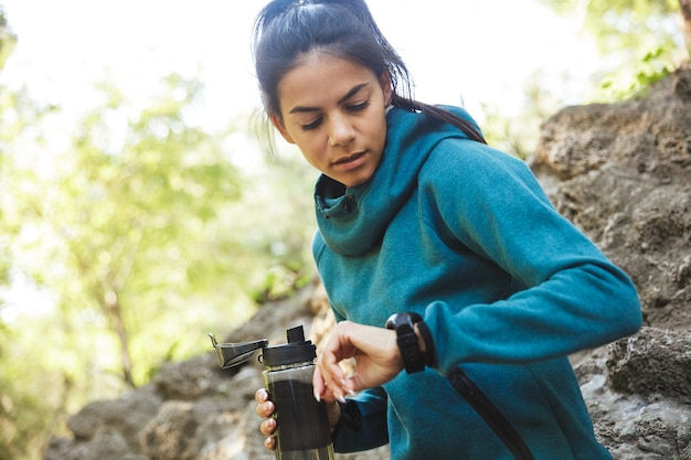 Gros plan d'une jolie jeune femme de remise en forme portant des vêtements de sport exerçant à l'extérieur, tenant une bouteille d'eau, en regardant sa montre