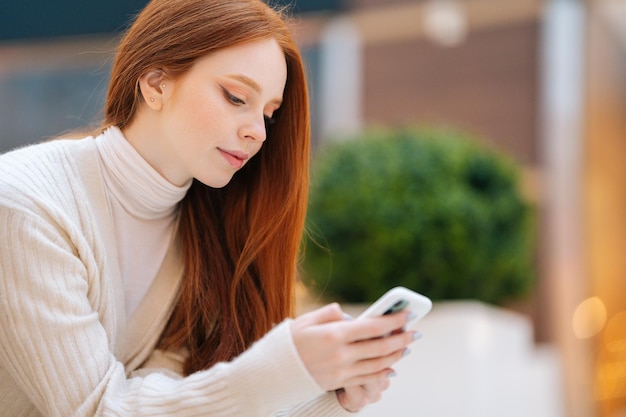 Gros plan d'une jolie jeune femme rousse utilisant un téléphone portable en tapant assis sur un banc au centre commercial avec un intérieur moderne, arrière-plan flou. Dame fatiguée se reposant après le shopping dans une boutique de vêtements.