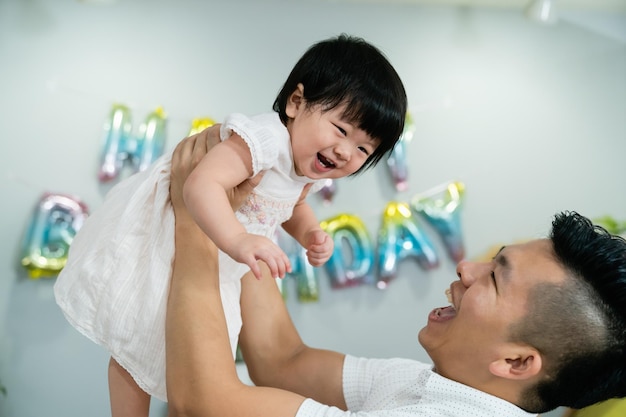 gros plan d'une jolie petite fille asiatique riant pendant que son père tient et joue avec elle contre un mur de fond blanc avec une décoration d'anniversaire