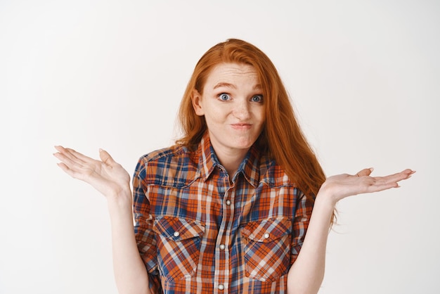 Photo gros plan d'une jolie rousse maladroite haussant les épaules en disant oups avec un visage coupable et un sourire confus debout sur fond blanc