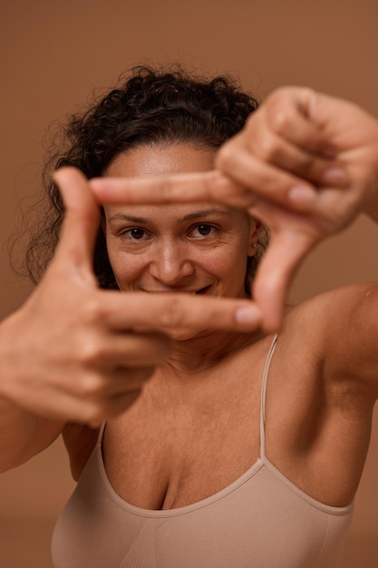 Photo gros plan d'une joyeuse femme bouclée aux cheveux noirs portant des sous-vêtements, sourire à pleines dents souriant regardant la caméra à travers un cadre de doigt, isolée sur fond beige avec espace de copie pour l'annonce