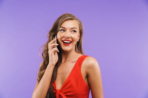 Gros plan de joyeuse femme excitée vêtue d'une robe rouge souriant et parlant au téléphone portable