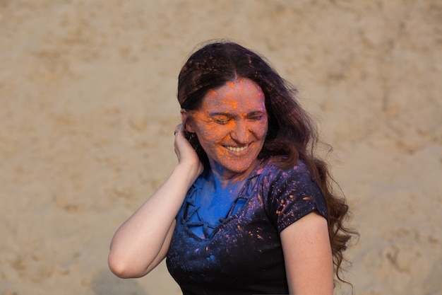 Gros plan d'un joyeux modèle brune aux longs cheveux bouclés recouverts de peinture sèche colorée Holi dans le désert