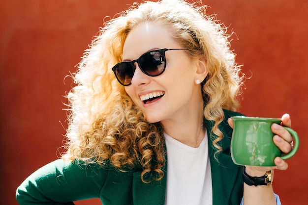 Gros plan latéral d'une femme séduisante portant des nuances élégantes et une veste tenant une tasse de thé verte ayant un sourire sincère sur son visage regardant de côté profiter de sa vie isolée sur fond orange