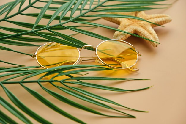 Gros plan sur des lunettes de soleil jaunes et des feuilles de palmier vue de dessus sur une surface beige