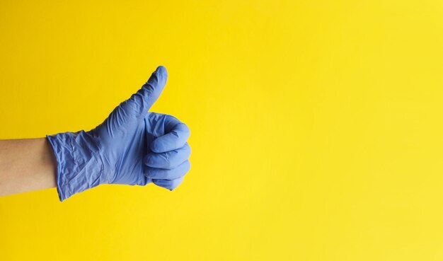 Gros plan de la main du médecin dans un gant bleu avec le pouce vers le haut sur fond jaune.