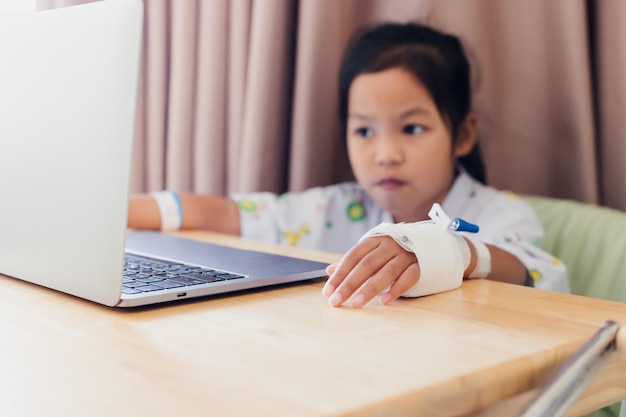 Gros plan sur la main d'un enfant malade qui a une solution IV pendant que l'enfant étudie un appel vidéo en ligne avec un ordinateur portable à l'hôpital. Isolement enseignement à domicile et enseignement à distance.