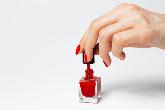 Gros plan d'une main féminine avec manucure rouge tenant un pinceau de vernis à ongles scintillant de couleur rouge sur fond blanc