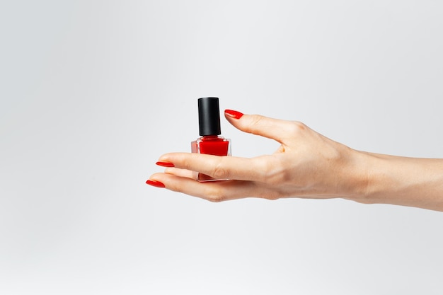 Photo gros plan d'une main féminine avec manucure rouge tenant un vernis à ongles scintillant de couleur rouge sur fond blanc