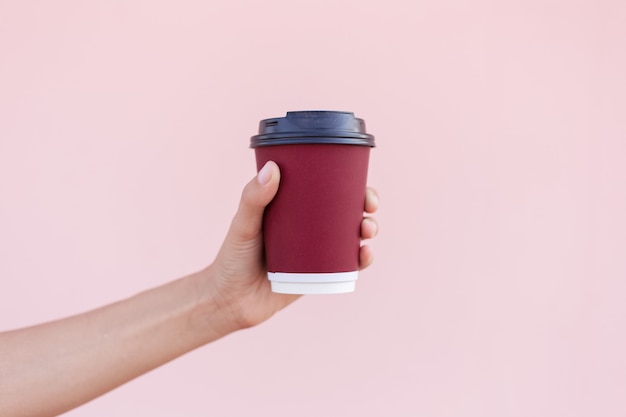 Gros plan d'une main féminine tenant une tasse de café en papier sur le fond de couleur rose pastel.