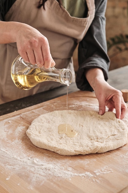 Gros plan d'une main féminine versant de l'huile du pichet sur la pâte tout en faisant une pizza maison