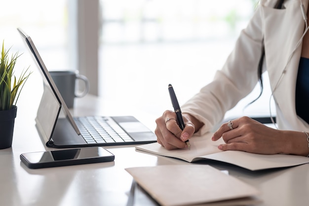 Gros plan d'une main de femme d'affaires tenant un stylo prenant des notes avec une tablette et un smartphone au bureau.