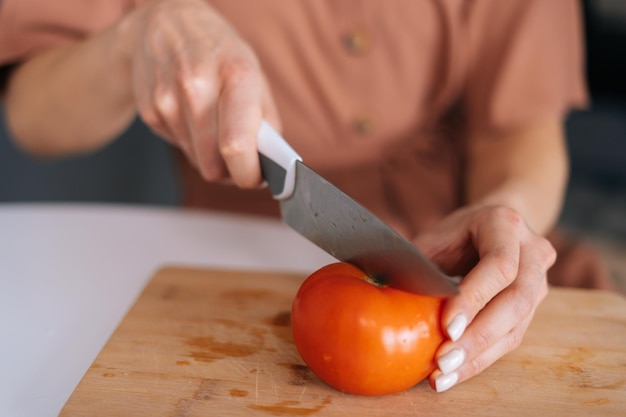Gros plan de la main d'une femme coupant des tomates fraîches à l'aide d'un couteau de cuisine sur une planche à découper en bois. Jeune femme coupant des tomates biologiques fraîches avec un couteau pour salade de légumes.