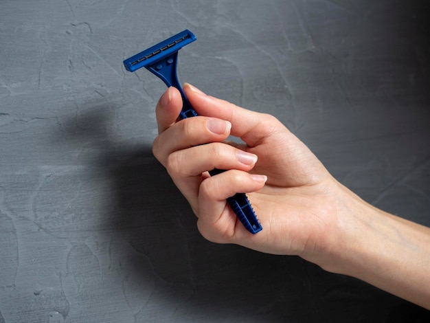 Gros plan de la main d'une femme tenant un rasoir en plastique bleu sur un fond texturé gris