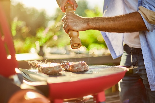 Gros plan sur la main de l'homme assaisonnant la viande sur le gril à gaz sur le barbecue en plein air