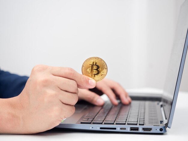 Gros plan de la main de l'homme tenant un bitcoin d'or et utilisant un ordinateur portable sur la table