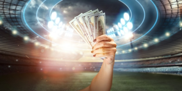Gros plan d'une main d'homme tenant des dollars américains dans le contexte du stade. Le concept des paris sportifs, faire un profit des paris, des jeux d'argent. Football américain.