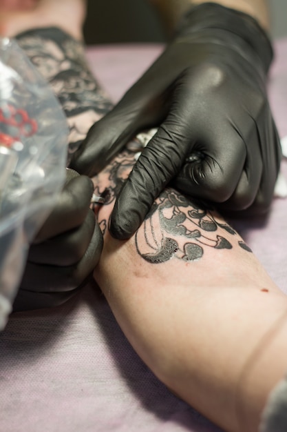 Gros plan de la main d'un maître de tatouage en gants noirs avec une machine à tatouer fait un tatouage selon le croquis