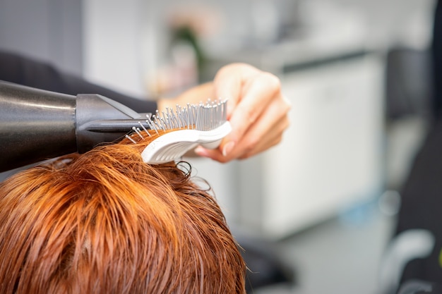 Gros plan de la main des maîtres avec brushing et brosse à cheveux soufflant les cheveux rouges des femmes dans un salon