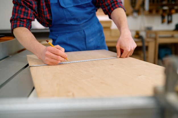 Gros plan de la main masculine mesurant la planche de bois. Charpentier homme utilisant un ruban à mesurer pour rendre les meubles vierges avant de travailler sur une scie circulaire à l'atelier