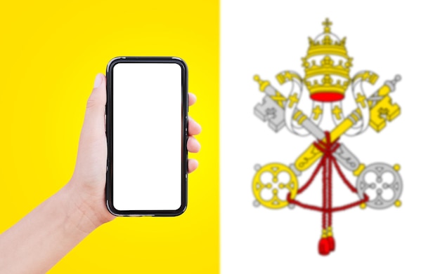 Gros plan d'une main masculine tenant un smartphone avec un blanc à l'écran sur fond de drapeau flou de la Cité du Vatican