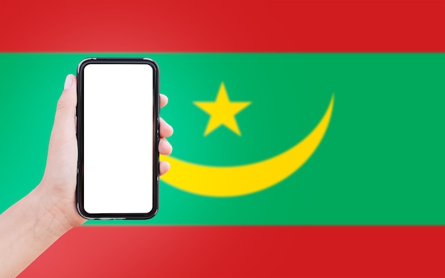 Gros plan sur une main masculine tenant un smartphone avec un blanc à l'écran sur fond de drapeau flou de la Mauritanie