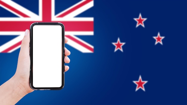 Gros plan d'une main masculine tenant un smartphone avec un blanc à l'écran sur fond de drapeau flou de la Nouvelle-Zélande