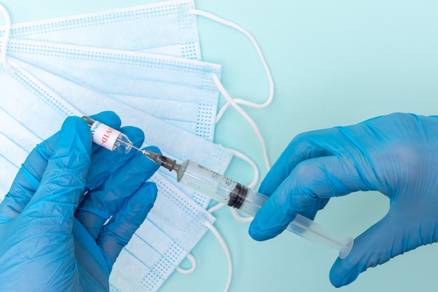 Gros plan d'une main de médecins dans des gants médicaux bleus en prenant une solution vaccinale dans une seringue sur un fond bleu avec des masques médicaux.