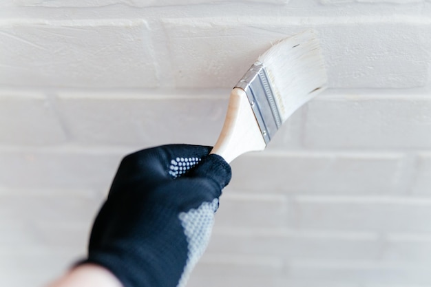 Gros plan de la main d'un travailleur avec un pinceau peignant un mur de briques avec de la peinture blanche