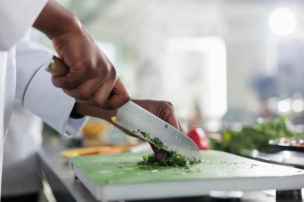 Gros plan sur des mains afro-américaines à l'aide d'un couteau professionnel pour hacher finement des herbes et des légumes biologiques frais pour la recette de ragoût. Cuire les verts de coupe pour un plat gastronomique servi au restaurant gastronomique.