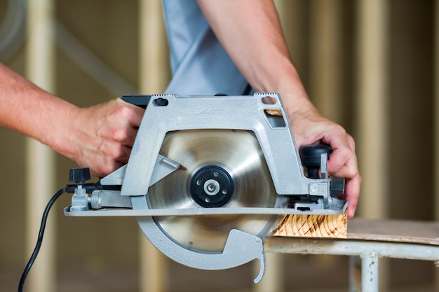 Gros plan des mains de charpentier musclé à l'aide de la nouvelle scie électrique circulaire forte et moderne pour couper la planche de bois dur. Outils professionnels pour la construction et le concept de construction.