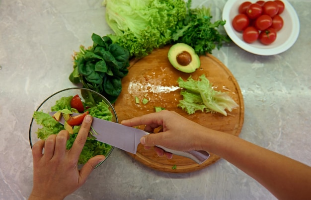 Gros plan sur les mains d'un chef féminin avec un couteau de cuisine coupant des tomates et les jetant dans un arc en verre transparent tout en préparant une salade végétalienne crue saine. Vue grand angle