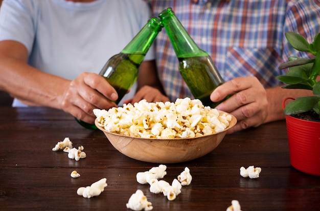 Gros plan sur les mains d'un couple senior caucasien portant un toast avec deux bouteilles de bière mangeant du pop-corn