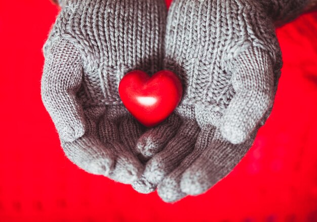 Gros plan des mains dans des mitaines tricotées grises tenant un coeur rouge