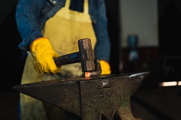 Gros plan des mains du forgeron travaillant avec un marteau sur un morceau de métal chaud.