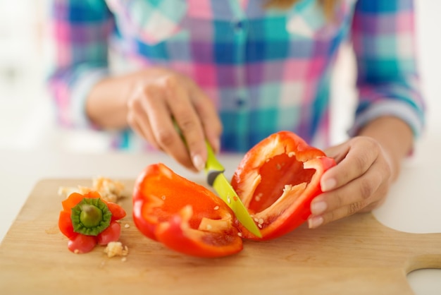 Gros plan sur des mains féminines coupant du paprika rouge sur le plateau de la cuisine.