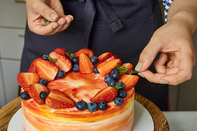 Gros plan sur des mains féminines décorant un gâteau fait maison avec des fraises et des bleuets. Boulangerie à domicile, petite entreprise.