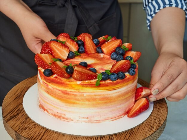 Gros plan sur des mains féminines décorant un gâteau fait maison avec des fraises et des bleuets. Boulangerie à domicile, petite entreprise.